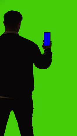 Vertikale-Videoaufnahme-Eines-Mannes,-Der-Ein-Mobiltelefon-Mit-Blauem-Bildschirm-Hält-Und-Von-Hinten-Als-Silhouette-Vor-Einem-Grünen-Bildschirm-Steht